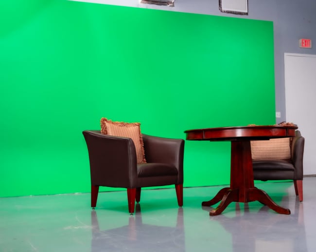 The image shows two dark leather chairs with decorative cushions positioned around a wooden table in front of a green screen.
