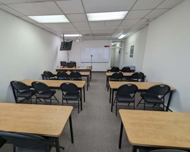 The image shows a classroom with rows of tables and chairs, a whiteboard, and a monitor at the front.