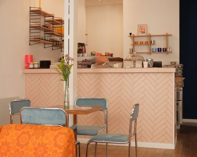 The image shows a cozy café interior with a seating area featuring a table and chairs, a counter displaying various food items, and decorative elements on the walls.