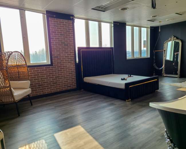 The room features a modern design with a bed, a vintage mirror, a freestanding bathtub, and a rattan chair against a brick wall.