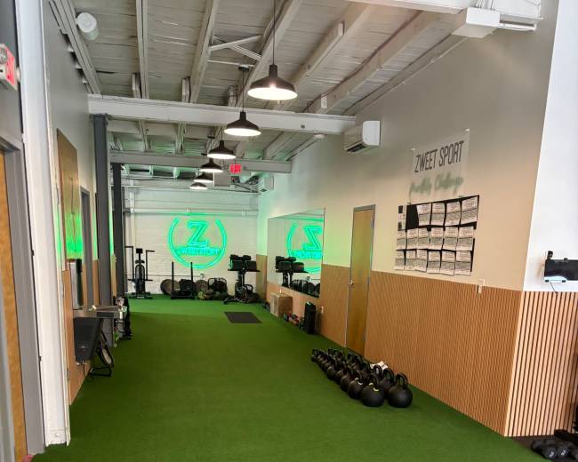 The image shows a gym interior with artificial grass flooring, workout equipment along the walls, and a glowing green logo prominently displayed.