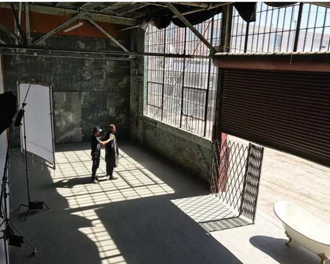 Two individuals stand in an industrial space with large windows and shadows cast on the floor, next to a freestanding bathtub.