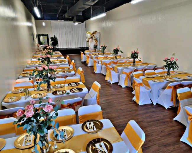 The image shows a banquet hall arranged with neatly set tables covered in white tablecloths and gold accents, adorned with floral centerpieces.