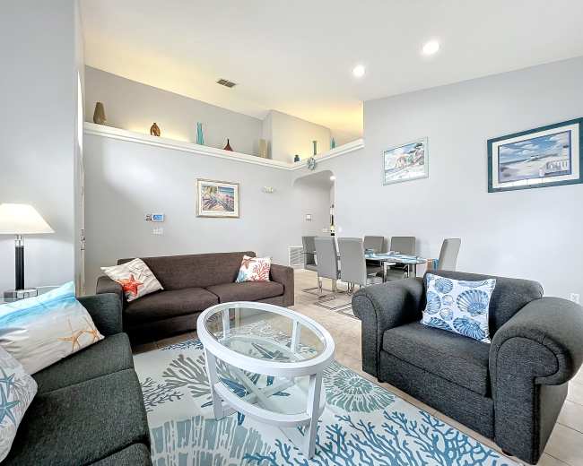 The image shows a modern living room featuring a sofa, armchairs, a glass coffee table, and decorative elements on the walls and shelves.