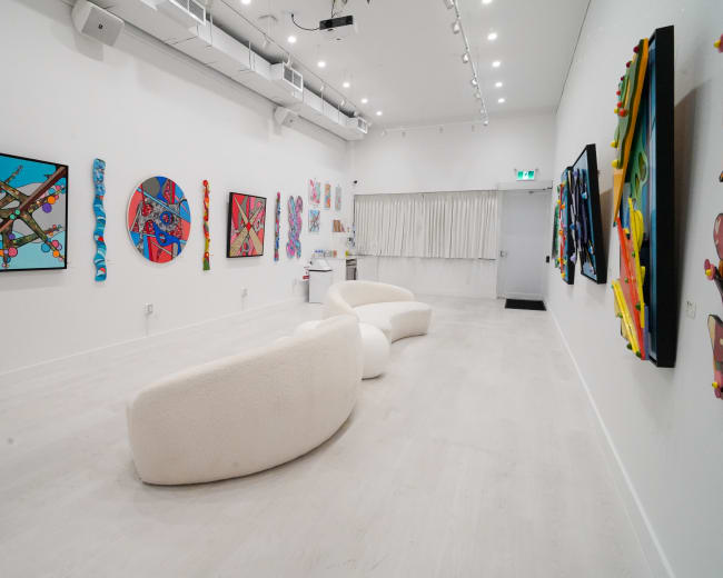 The image features a modern art gallery with colorful abstract paintings displayed on white walls and a curved white seating area in the center.