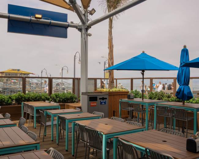 The image shows an outdoor dining area with blue umbrellas and wooden tables overlooking a harbor with boats in the background.