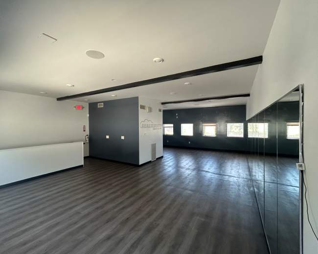 The image shows a spacious, modern room with gray walls, wooden beams, and large windows, complemented by a mirrored wall on one side.