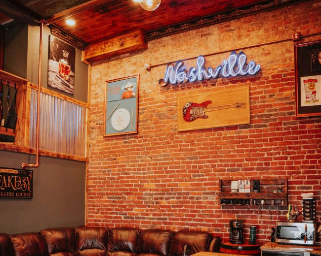 The image shows a cozy lounge area featuring a leather sofa, a kitchen island with a granite countertop, and a brick wall adorned with a neon sign reading "Nashville."