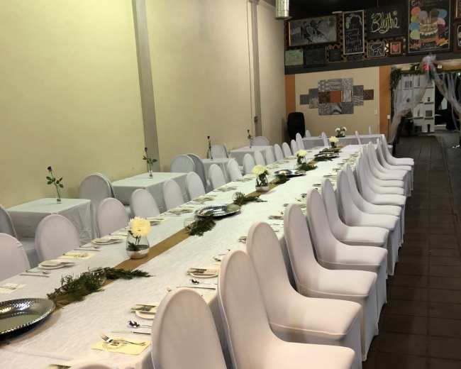 A long banquet table is set up with white chairs and decorated with simple centerpieces in a spacious indoor venue.