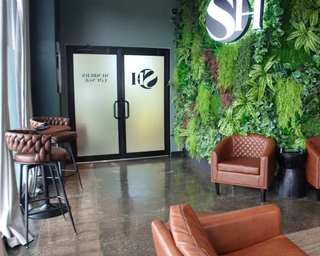 The image shows a modern lounge area with two brown leather chairs and a green plant wall, featuring a large logo on the wall and a glass door marked with a name.