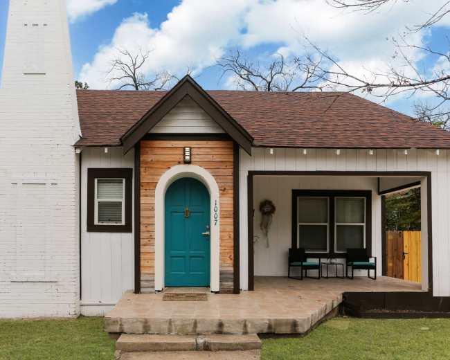 The image shows a single-story house with a white exterior, a wooden accent wall, a teal front door, and a small porch with two chairs.
