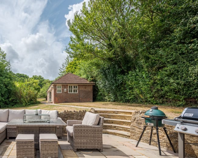 A patio area features a large seating arrangement with a glass table, surrounded by trees and a building in the background.