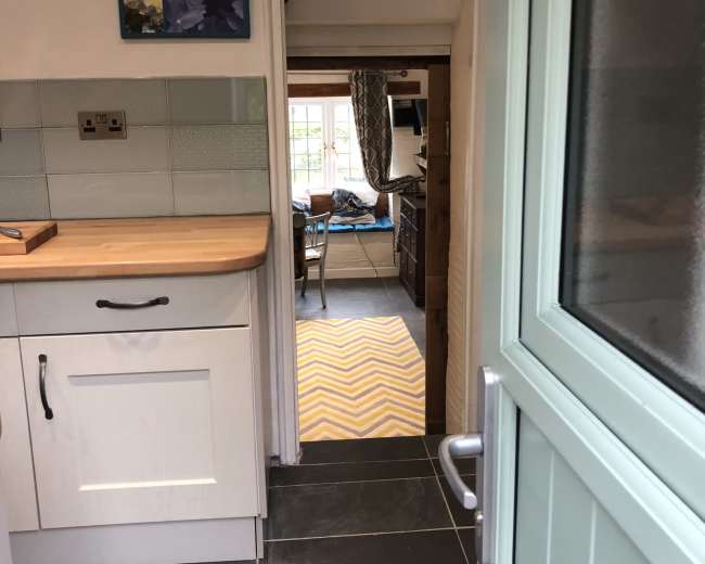 A door opens from a kitchen with a wooden countertop, leading to a hallway and visible room beyond with a patterned rug.