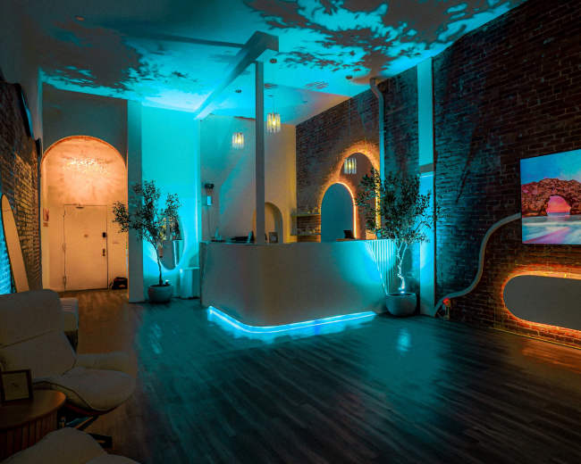 A modern lounge features exposed brick walls, ambient blue lighting, and a large screen displaying a sunset over a beach.