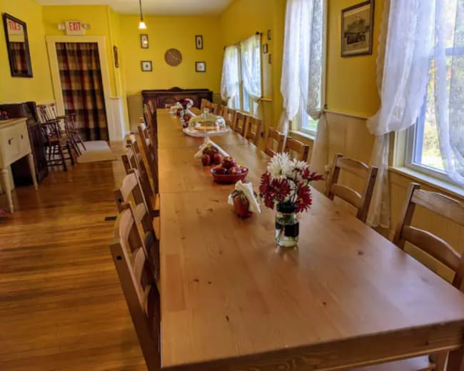 A long wooden dining table is set with fruit and flowers in a brightly lit room with yellow walls and large windows.