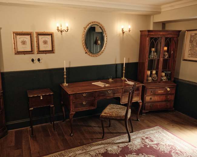 The image depicts a historical-style office with wooden furniture, including a desk and shelves, adorned with decorative items and framed pictures on the walls.