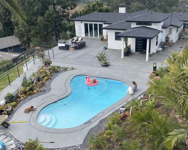 A person sits by a swimming pool shaped like a large kidney, while another swims with a pink float, surrounded by a landscaped yard and a modern house.