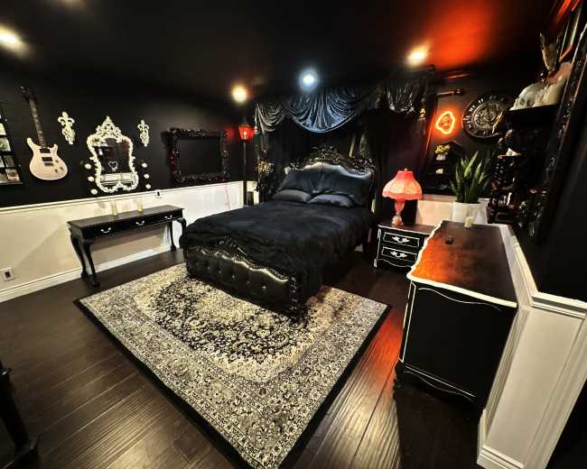 The image depicts a dark-themed bedroom featuring a black ornate bed with a decorative headboard, a patterned rug, and various furnishings including a dresser and side tables.