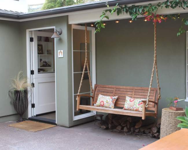 A swing bench with decorative cushions hangs on a porch beside a green wall and large windows.