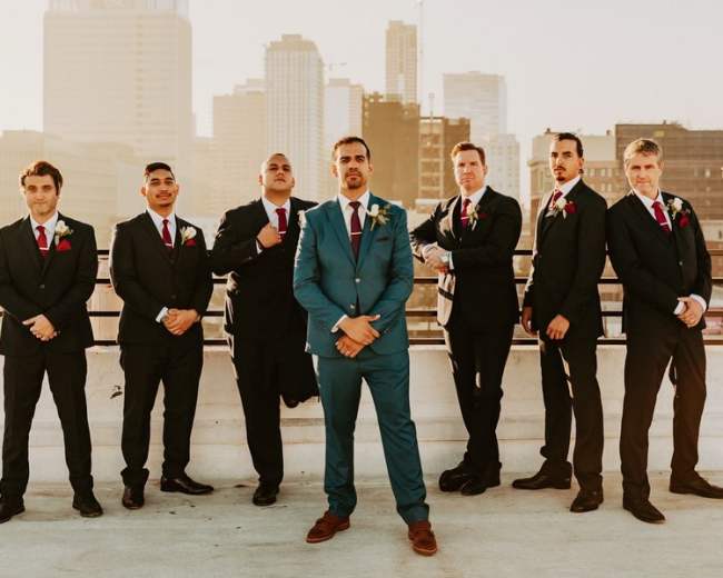 A group of seven men dressed in suits stands on a rooftop with a city skyline in the background.