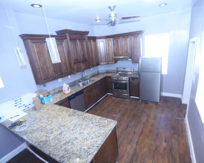 The image shows a kitchen with dark wooden cabinets, granite countertops, a stainless steel refrigerator, and an oven, featuring a wooden floor and a ceiling fan.