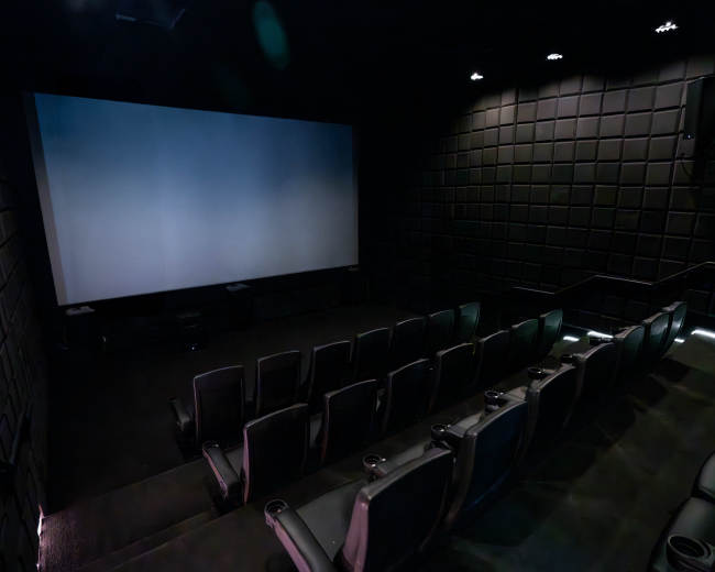 The image shows a darkened movie theater with a large blank screen and rows of empty reclining seats.
