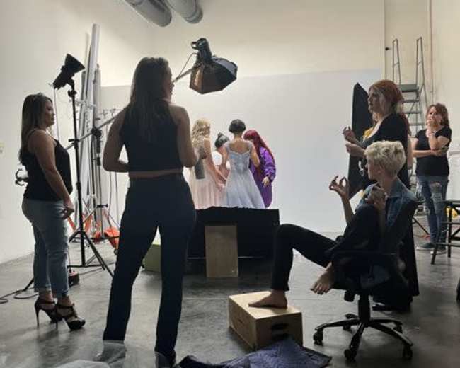 A group of people works in a studio setting, with some preparing for a photo shoot while others pose in elegant dresses.