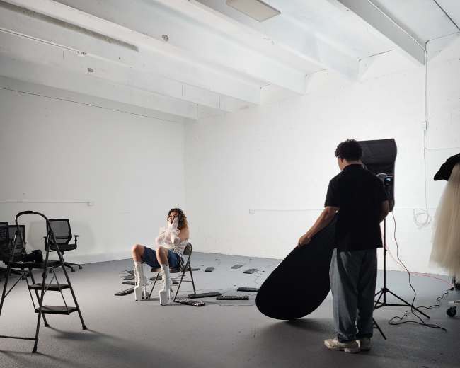 A photographer adjusts a reflector while a model sits on a chair in a sparsely furnished studio with bright lighting.