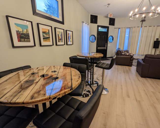 A modern interior space with two wooden tables surrounded by black chairs, framed artwork on the walls, and a seating area with a brown couch and curtains.