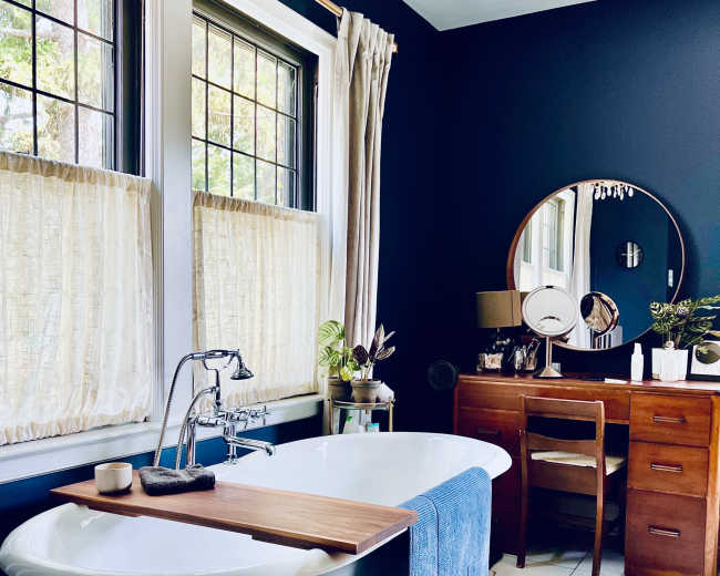 A bathroom featuring a freestanding tub with a wooden side table, a retro vanity with a round mirror, and large windows covered by light curtains.