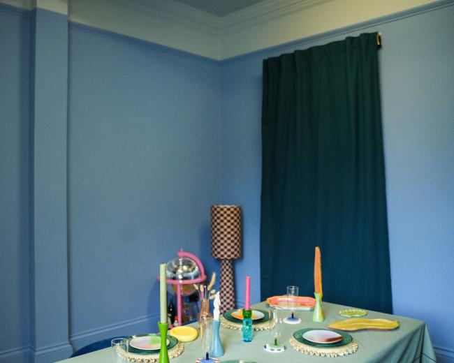 A dining table is set with colorful plates and candles in a room with blue walls and a green curtain.