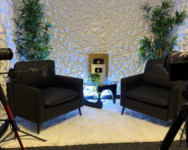The image shows a modern interview setup with two black chairs, a decorative wall, plants, and a golden YouTube play button placed on a small table.