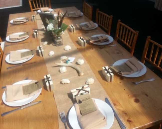 A long wooden table is set with neatly arranged plates, utensils, and a burlap runner, all under a tent with a polished floor in the background.