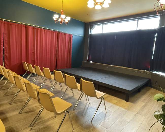 The image shows a small theater space with a raised stage and rows of plastic chairs arranged facing the stage, framed by red and dark curtains.