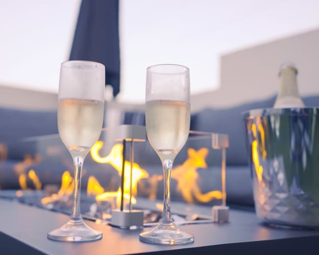 Two glasses of champagne sit beside a fire feature on a table, with a bottle of champagne in an ice bucket nearby, set on a patio.