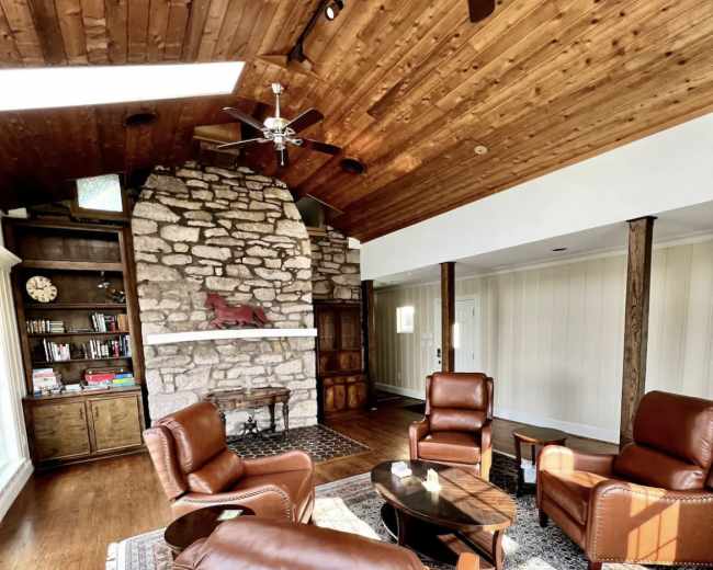 A cozy living room featuring a stone fireplace, wooden ceiling, and four brown leather chairs arranged around a circular table on a patterned rug.