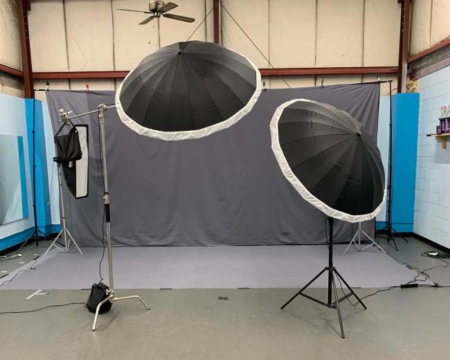 A photography studio setup with two large umbrella softboxes positioned in front of a gray backdrop on a flat floor.