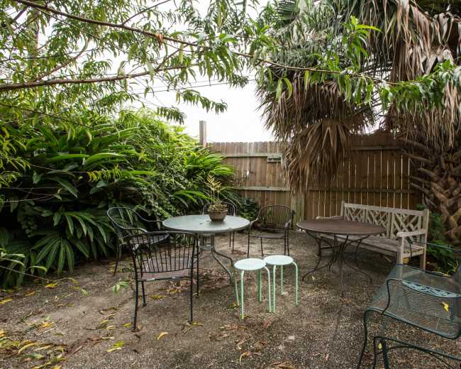A small patio area features a round table with chairs surrounded by lush greenery and a wooden fence.
