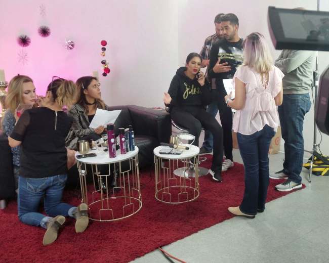 A group of people engages in conversation on a set with cameras, located in a room featuring a red carpet and decorative backdrop.