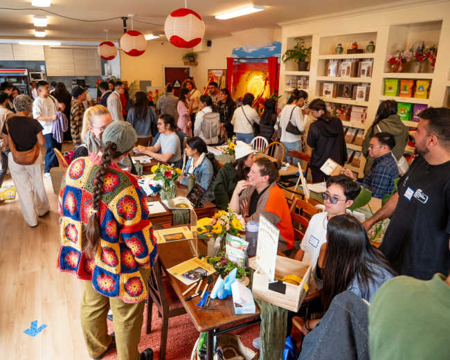 A large group of people gathers in a brightly lit community space filled with tables, decorations, and a lively atmosphere.