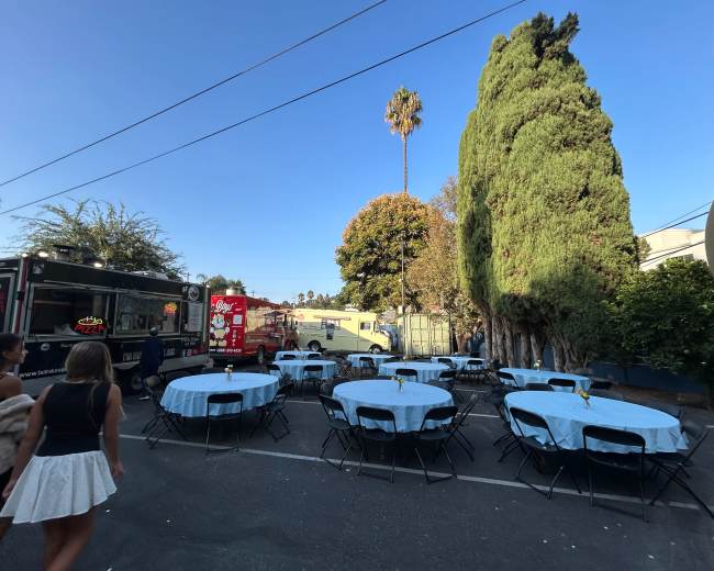 The scene features an outdoor event space with tables covered in light blue tablecloths and food trucks parked nearby, surrounded by palm trees and greenery.