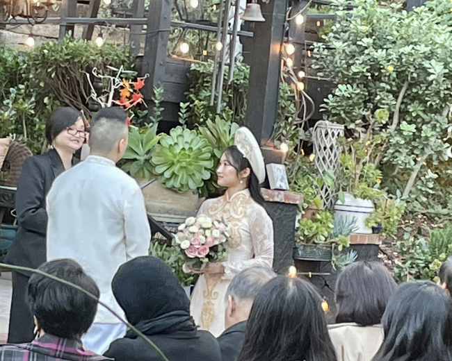 A bride in a traditional dress holds a bouquet while standing with a groom during a wedding ceremony outdoors, surrounded by greenery and string lights.