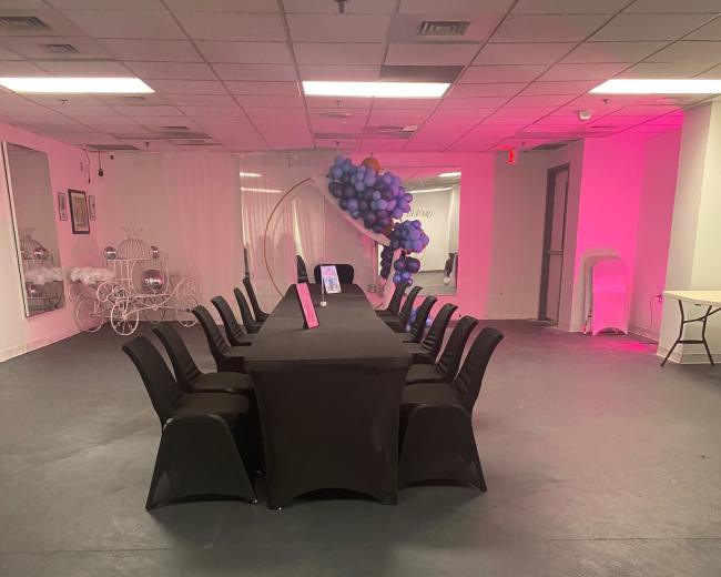 A long table with black tablecloths and chairs is set up in an empty room with pink lighting and decorative elements in the background.