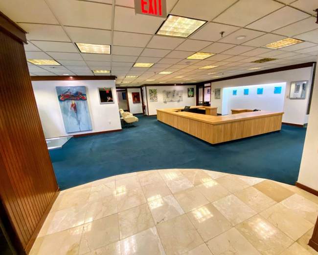 The image shows a spacious office area with a wooden reception desk, carpeted flooring, and artwork displayed on the walls.