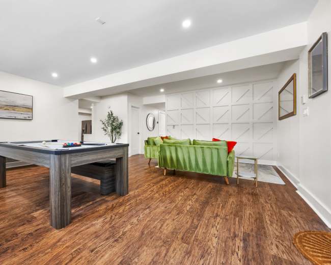 The image shows a spacious living area featuring a billiards table, a green couch, and wooden flooring.