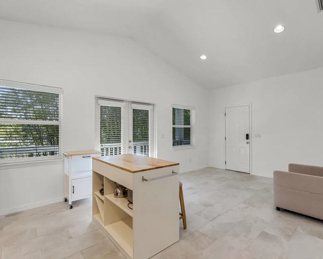 The image shows a bright, open living space featuring a small kitchen island, a couch, and large windows.