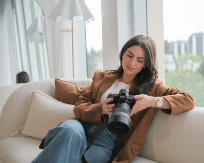 A woman is sitting on a sofa, holding a camera while looking at it thoughtfully.