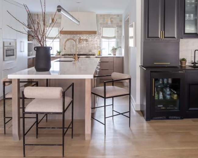 A modern kitchen features a large island with bar stools, a cabinetry section with glass doors, and a high ceiling with exposed beams.