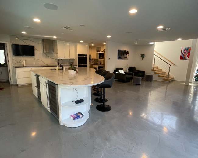 A modern kitchen with a large island, sleek countertops, and an open living area featuring a staircase and seating.