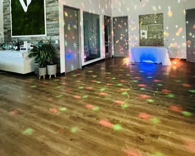 The image shows an indoor venue with a wooden floor, colorful lights reflecting off the walls and ceiling, and a bar area in the background.
